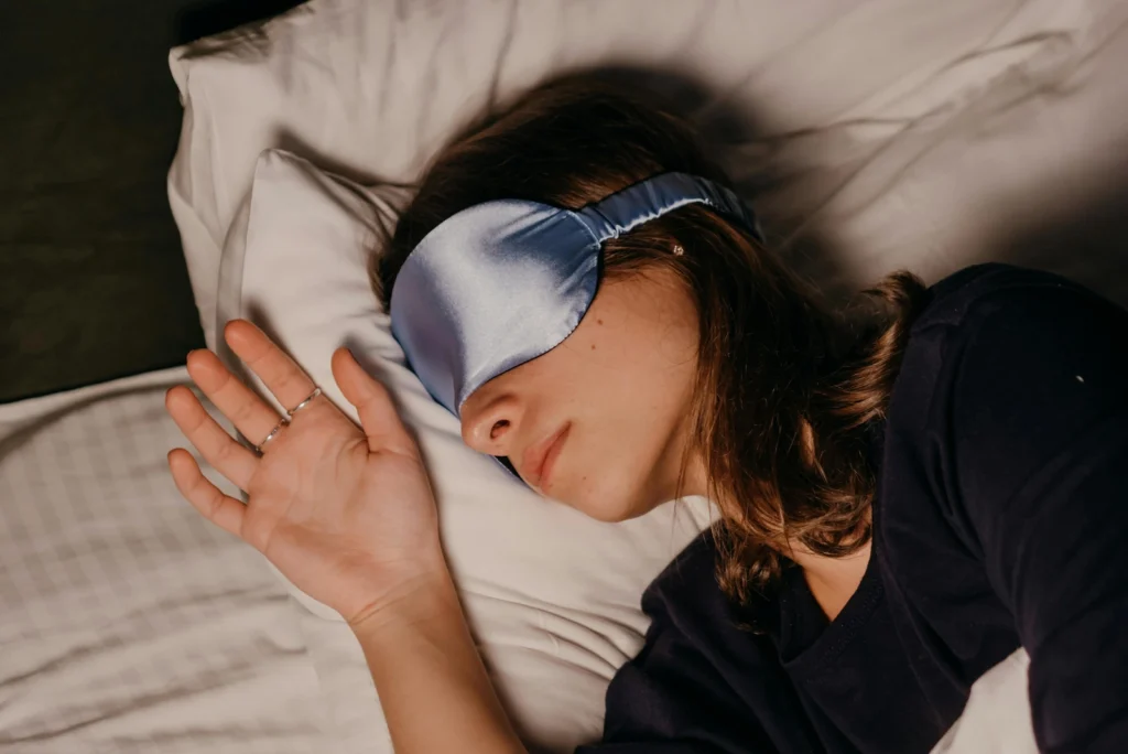 Close-up of a young woman with a silk eye mask sleeping peacefully on a white pillow, highlighting the essential need for quality sleep for brain function and memory consolidation.