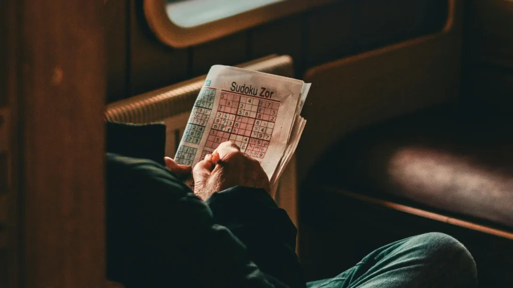 Person solving a Sudoku puzzle on a newspaper while sitting in a cozy, dimly lit train compartment
