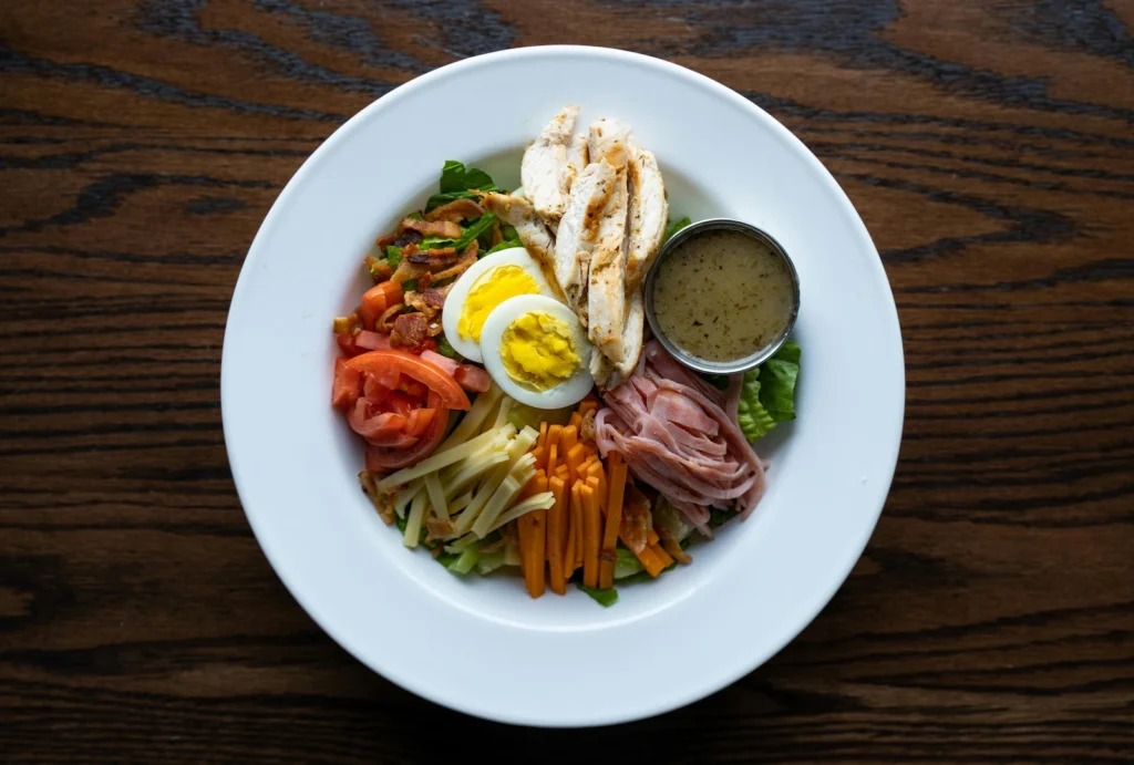 A colorful Cobb salad served in a white bowl, containing sliced chicken, ham, boiled egg halves, bacon bits, cheese, tomatoes, and lettuce, which represents excellent foods for brain function.