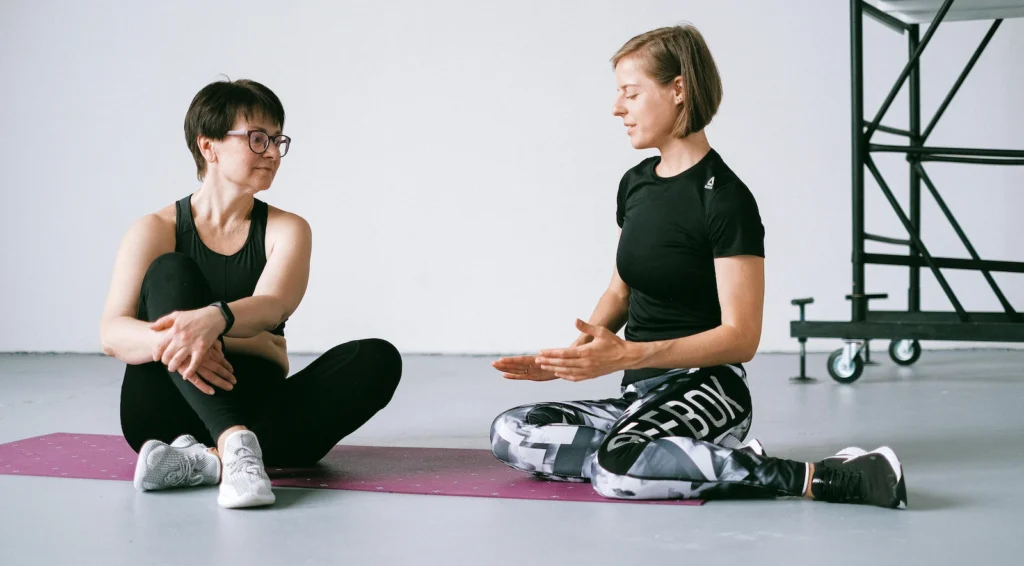 Two middle-aged women sitting on a yoga mat in a gym, discussing exercise routine which is important for Midlife brain health.